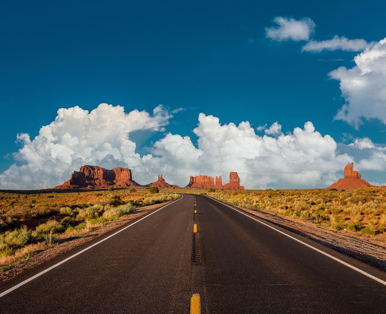 Monument valley, blue sky and clouds with a road heading straight fading in the distance with desert mountains in arizona